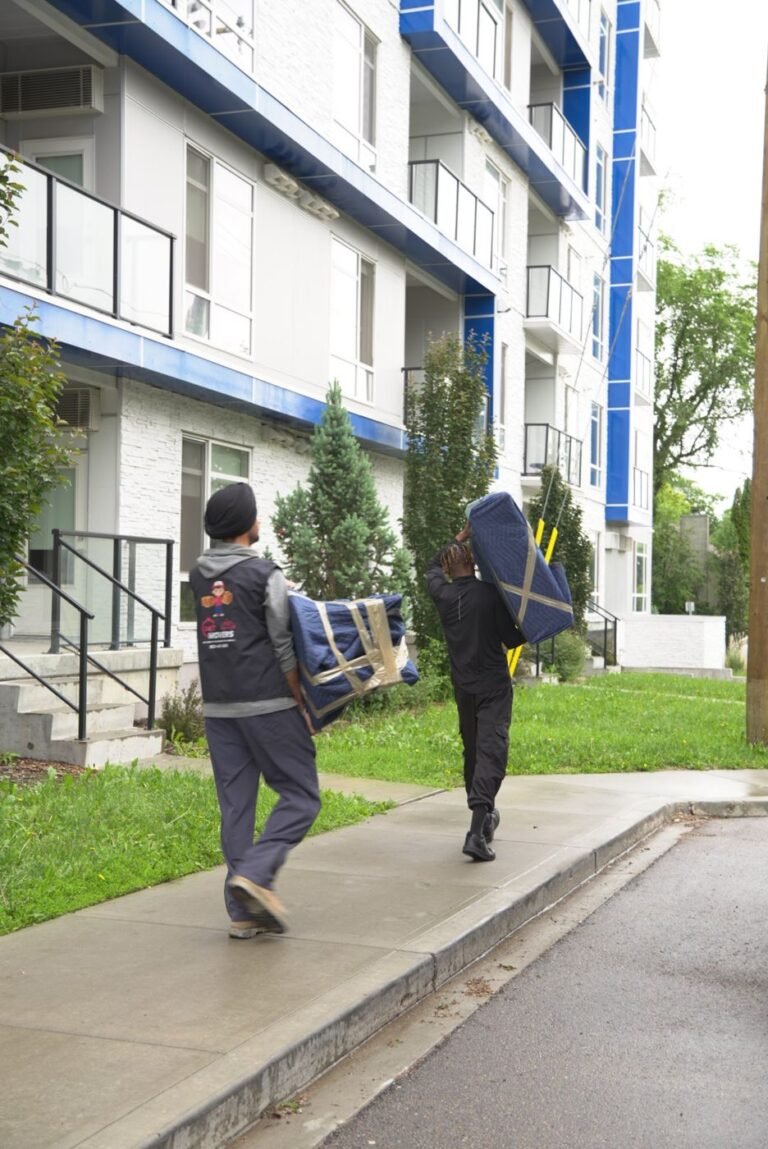 Two movers carrying blanket-wrapped furniture to an apartment building